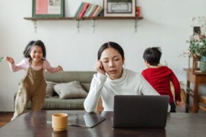 Mom slumped over her computer looking exhausted with a cup of coffee and her two children a boy about 10 years old and a girl about 8 years old are running around behind her. 